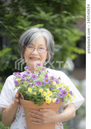 一位微笑的白髮老婦人喜歡園藝，她正在照顧院子裡的花壇和植物，目光直視鏡頭。 140540654