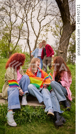 Three young women sitting with rainbow flags and two men walking to them in green park 140540711