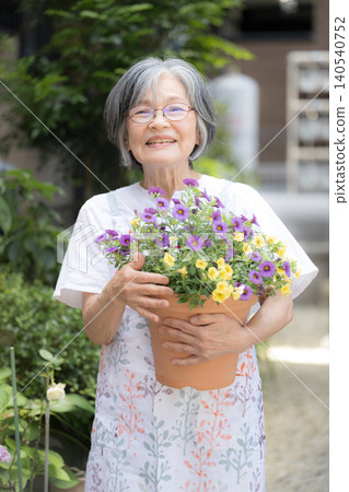 一位微笑的白髮老婦人喜歡園藝,她正在照顧院子裡的花壇和植物,目光直視鏡頭。 一位微笑的白髮老婦人喜歡園藝,她正在照顧院子裡的花壇和植物,目光直視鏡頭。 140540752
