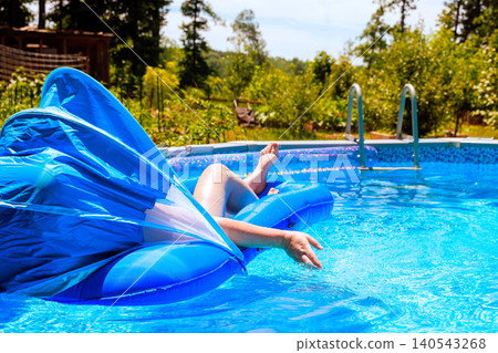 Woman relaxes on blue raft in pool while enjoying sunny day surrounded by trees, greenery. 140543268