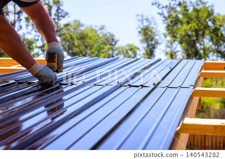 Roofing builder secures metal roof panels with power drill tool while working on construction site. Roofing builder secures metal roof panels with power drill tool while working on construction site. 140543282