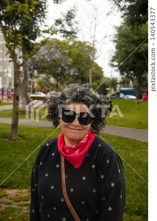 A woman wearing sunglasses and a red bandana is smiling in a park 140543377