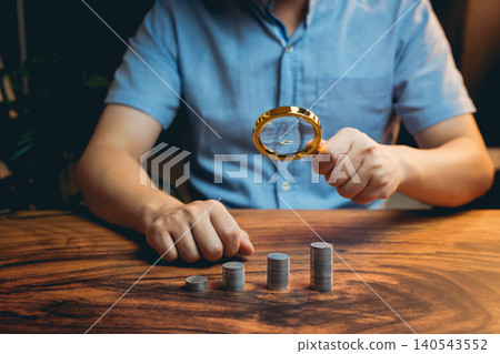 Hand Holding Magnifying Glass over Stacked Coins on Wooden Table with Soft Background for Financial Analysis Hand Holding Magnifying Glass over Stacked Coins on Wooden Table with Soft Background for Financial Analysis 140543552