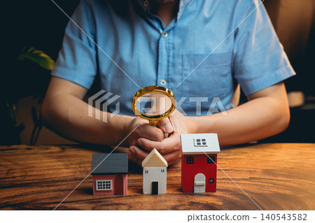 Person Examining Miniature Houses with Magnifying Glass on Wooden Table for Real Estate and Investment Concepts Person Examining Miniature Houses with Magnifying Glass on Wooden Table for Real Estate and Investment Concepts 140543582