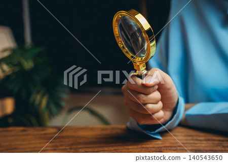 Detailed Close-Up of Hand Holding Gold Magnifying Glass Over Wooden Surface in Office Environment 140543650