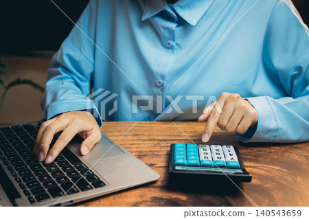 Professional User Working on Laptop While Using Calculator for Financial Calculations in Modern Workspace Professional User Working on Laptop While Using Calculator for Financial Calculations in Modern Workspace 140543659