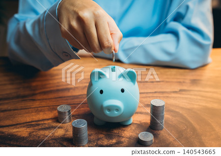 Hand Inserting Coin Into Blue Piggy Bank on Wooden Table Surrounded by Stacks of Coins in Soft Lighting Hand Inserting Coin Into Blue Piggy Bank on Wooden Table Surrounded by Stacks of Coins in Soft Lighting 140543665
