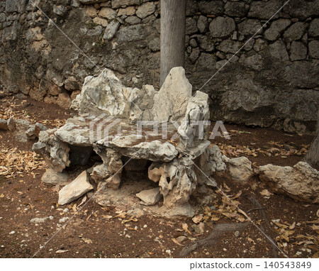 A weathered garden bench built from natural rocks stands against an ancient stone wall. Leaves cover the ground nearby in this quiet corner of the garden. 140543849