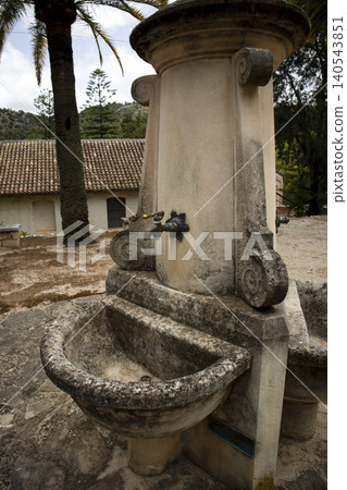 A vintage drinking fountain stands in a quiet area. The fountain has a brass tap and stone basins, surrounded by Mediterranean buildings and palm trees. Many people visit this location. A vintage drinking fountain stands in a quiet area. The fountain has a brass tap and stone basins, surrounded by Mediterranean buildings and palm trees. Many people visit this location. 140543851