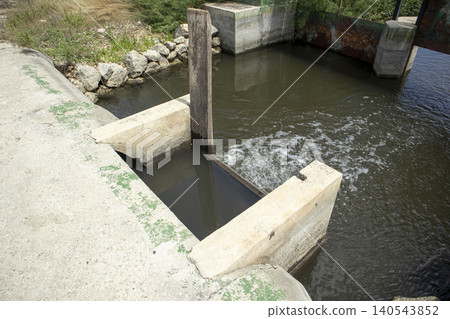 Water moves steadily through a concrete canal system in the countryside. An old wooden sluice gate controls the flow of water. This scene captures rural life in a rice farming area. Water moves steadily through a concrete canal system in the countryside. An old wooden sluice gate controls the flow of water. This scene captures rural life in a rice farming area. 140543852