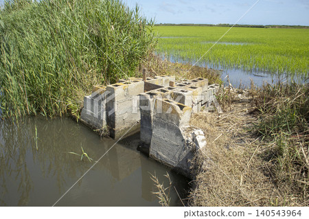 The photo shows a cinder block water control structure in a natural reserve. It regulates water flow for rice paddies, surrounded by green fields and a water body. The photo shows a cinder block water control structure in a natural reserve. It regulates water flow for rice paddies, surrounded by green fields and a water body. 140543964
