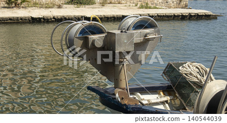 A hydraulic net hauler drum is seen on the bow of a fishing boat. The boat is docked in a port. The water reflects the sunlight and shows a calm harbor environment. 140544039