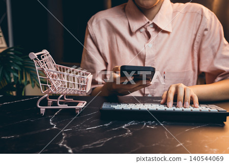 Young person shopping online with smartphone and keyboard, small shopping cart on marble table, modern lifestyle 140544069