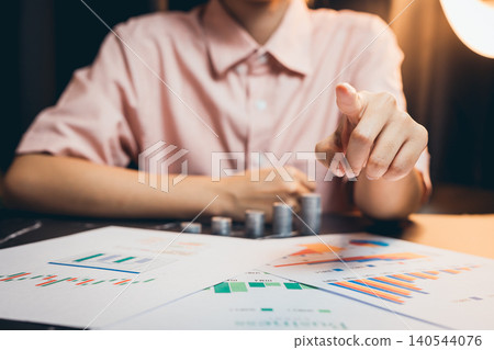 Businessperson Analyzing Data Trends and Giving Instructions with Graphs and Charts on a Desk in a Dimly Lit Office 140544076
