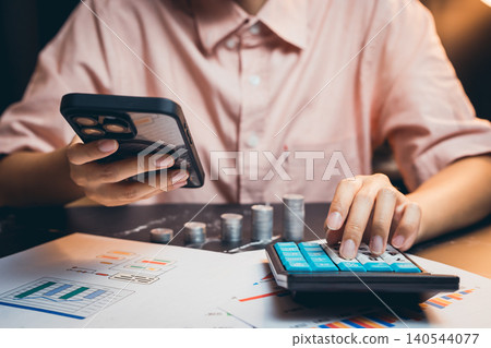 Person in Pink Shirt Using Smartphone and Calculator for Financial Analysis with Charts and Coins on Desk 140544077