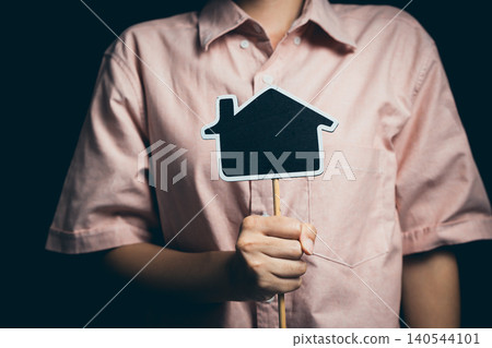 Person Holding Paper House Icon on Stick in Pink Shirt Against Dark Background for Real Estate Concept Person Holding Paper House Icon on Stick in Pink Shirt Against Dark Background for Real Estate Concept 140544101