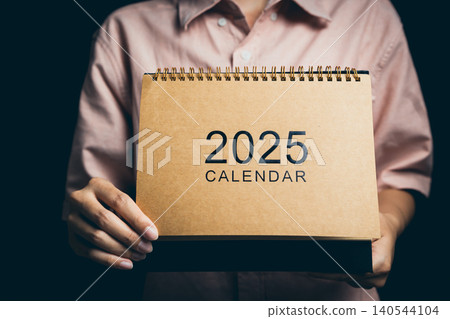 Person Holding a 2025 Calendar in a Neutral Setting with Soft Lighting and a Focus on Time Management Person Holding a 2025 Calendar in a Neutral Setting with Soft Lighting and a Focus on Time Management 140544104