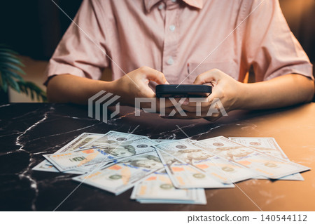 Young person using smartphone with cash spread on table, financial transaction, banking technology concept Young person using smartphone with cash spread on table, financial transaction, banking technology concept 140544112