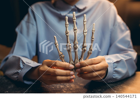 Halloween prop concept. Human skeletal hand model in a person's hands showcasing anatomy in a dimly lit environment Halloween prop concept. Human skeletal hand model in a person's hands showcasing anatomy in a dimly lit environment 140544171