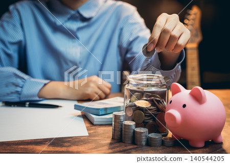 Business concept. Person Placing Coin in Jar Next to Piggy Bank and Stack of Coins on Wooden Table with Notebook in Background Business concept. Person Placing Coin in Jar Next to Piggy Bank and Stack of Coins on Wooden Table with Notebook in Background 140544205