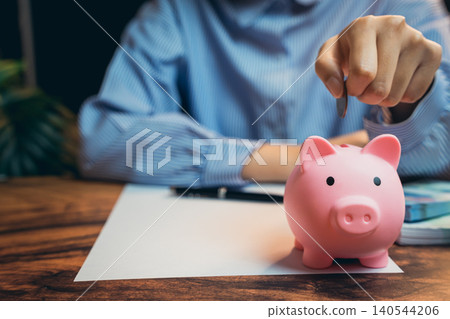 Business concept. Person Saving Money by Inserting Coin into Pink Piggy Bank on Wooden Table with Financial Documents Nearby Business concept. Person Saving Money by Inserting Coin into Pink Piggy Bank on Wooden Table with Financial Documents Nearby 140544206