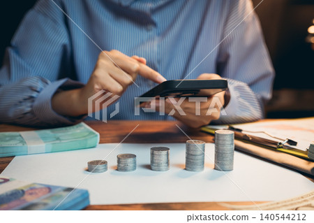 Business concept. Person Analyzing Financial Data Using Calculator with Stacked Coins and Banknotes on Wooden Desk Business concept. Person Analyzing Financial Data Using Calculator with Stacked Coins and Banknotes on Wooden Desk 140544212