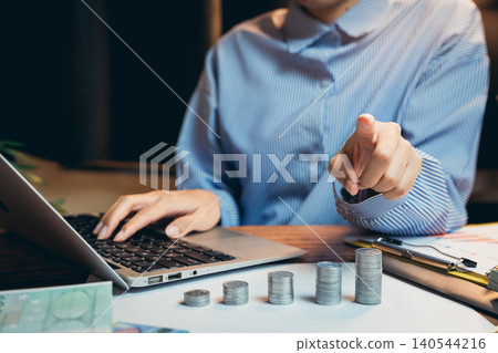 Business concept. Businessperson Pointing at Stacked Coins while Working on Laptop in Modern Office Environment 140544216