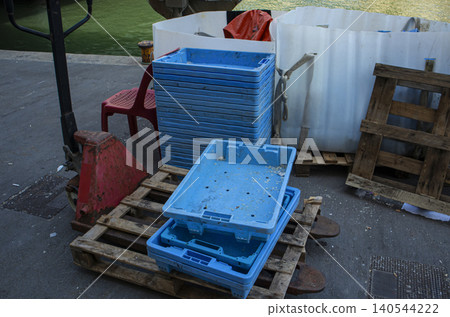 Blue plastic fish crates are stacked on a wooden pallet on a concrete pier. A pallet jack is nearby. This scene shows work in a busy harbor for commercial fishing. 140544222