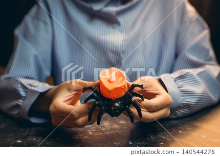 Halloween prop concept. Hands Holding a Black Spider Toy with a Halloween Orange Pumpkin on a Dark Surface in a Cozy Indoor Setting Halloween prop concept. Hands Holding a Black Spider Toy with a Halloween Orange Pumpkin on a Dark Surface in a Cozy Indoor Setting 140544278