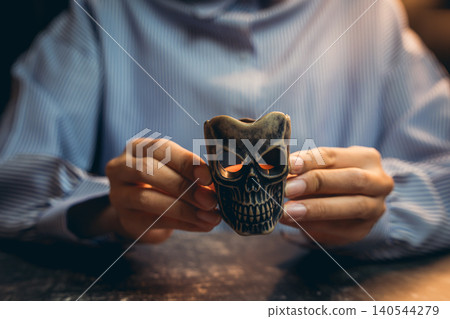 Halloween prop concept. Hands Holding Decorative Skull Figurine with Glowing Eyes in Dimly Lit Room Halloween prop concept. Hands Holding Decorative Skull Figurine with Glowing Eyes in Dimly Lit Room 140544279