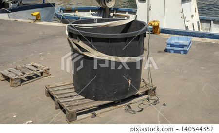 A large black plastic container is placed on a wooden pallet at a commercial shipping dock. It is secured with cargo straps. The harbor is busy with fishing boats nearby. A large black plastic container is placed on a wooden pallet at a commercial shipping dock. It is secured with cargo straps. The harbor is busy with fishing boats nearby. 140544352