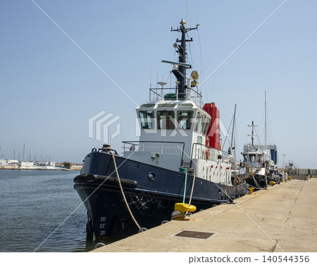 The tugboat is securely moored at a concrete pier in the harbor. It is prepared for operations under bright sunlight. The surroundings show other vessels and calm water. The tugboat is securely moored at a concrete pier in the harbor. It is prepared for operations under bright sunlight. The surroundings show other vessels and calm water. 140544356
