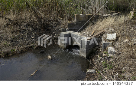 A rustic concrete culvert releases water into a drainage ditch in an ecological farming area. The setting shows rural infrastructure at work during daytime. A rustic concrete culvert releases water into a drainage ditch in an ecological farming area. The setting shows rural infrastructure at work during daytime. 140544357