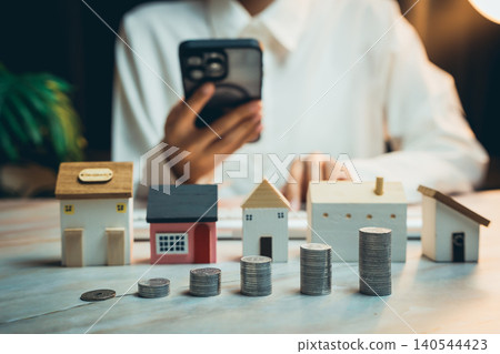 Home buying concept. Person Calculating Real Estate Investment Costs with Toy Houses and Coins on a Table in a Bright Environment Home buying concept. Person Calculating Real Estate Investment Costs with Toy Houses and Coins on a Table in a Bright Environment 140544423