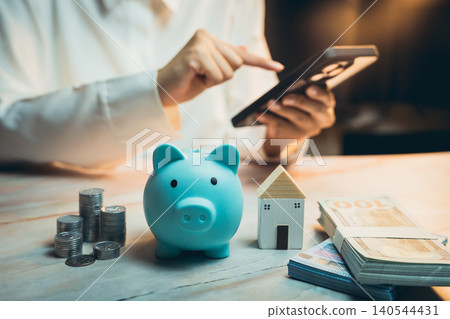 Home buying concept. Person Using Smartphone for Financial Management with Piggy Bank, House Model, Coins, and Banknotes on Wooden Table Home buying concept. Person Using Smartphone for Financial Management with Piggy Bank, House Model, Coins, and Banknotes on Wooden Table 140544431