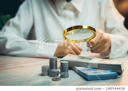 Business concept. Person using magnifying glass to examine cash and coins on table in indoor setting with soft lighting Business concept. Person using magnifying glass to examine cash and coins on table in indoor setting with soft lighting 140544434