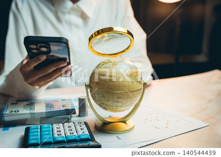 Business concept. Person Using Smartphone with Globe, Calculator, Cash, and Charts on Desk in Modern Office Setting Business concept. Person Using Smartphone with Globe, Calculator, Cash, and Charts on Desk in Modern Office Setting 140544439
