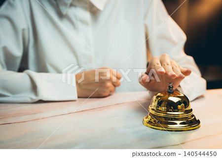 Business concept. Hand Reaching for a Golden Reception Bell on a Wooden Counter in a Warmly Lit Environment Business concept. Hand Reaching for a Golden Reception Bell on a Wooden Counter in a Warmly Lit Environment 140544450
