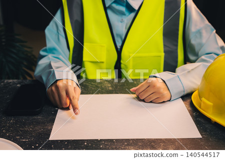 Engineer concept. Construction Worker in Safety Vest Pointing at Blank Sheet of Paper on Table with Hard Hat and Smartphone Nearby 140544517