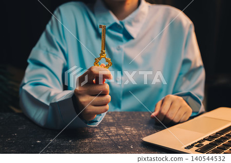 Success key concept. Golden Key Held by Person in Blue Shirt Next to Laptop in Dimly Lit Environment Success key concept. Golden Key Held by Person in Blue Shirt Next to Laptop in Dimly Lit Environment 140544532