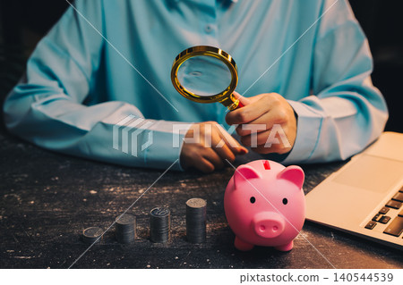 Saving concept. Person in Blue Shirt Using Magnifying Glass to Inspect Piggy Bank with Coins on Table Next to Laptop Saving concept. Person in Blue Shirt Using Magnifying Glass to Inspect Piggy Bank with Coins on Table Next to Laptop 140544539