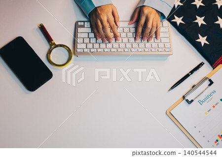 Business concept. Hands Typing on Keyboard with American Flag, Magnifying Glass, Phone, and Clipboard on White Desk Surface Business concept. Hands Typing on Keyboard with American Flag, Magnifying Glass, Phone, and Clipboard on White Desk Surface 140544544