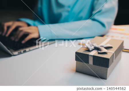 Gift concept. Gift box on table next to person typing on laptop computer, celebrating special occasions and festive moments Gift concept. Gift box on table next to person typing on laptop computer, celebrating special occasions and festive moments 140544562
