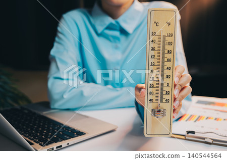Thermometer concept. Person Holding Vintage Thermometer While Working on Laptop in Cozy Indoor Environment Thermometer concept. Person Holding Vintage Thermometer While Working on Laptop in Cozy Indoor Environment 140544564