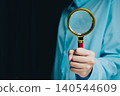 Businessman Holding Magnifying Glass in Blue Shirt with Dark Background for Inspection and Investigation Purposes 140544609