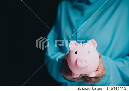 Businessman Holding Pink Piggy Bank Against Dark Background for Saving Money and Financial Planning Concept Businessman Holding Pink Piggy Bank Against Dark Background for Saving Money and Financial Planning Concept 140544610