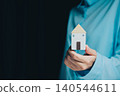 Businessman Holding Small Model House with Blue Shirt in Dark Background Representing Home and Real Estate Concept 140544611