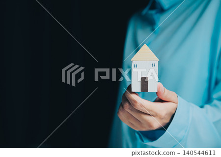 Businessman Holding Small Model House with Blue Shirt in Dark Background Representing Home and Real Estate Concept Businessman Holding Small Model House with Blue Shirt in Dark Background Representing Home and Real Estate Concept 140544611