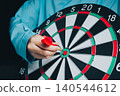 Business Target concept. Person wearing blue shirt aiming dart towards bullseye on dartboard with focus and precision in a competitive game 140544612