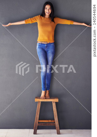 Woman, confident and stand on chair in portrait, smile and fashion on wall background. Female person, clothes and happy lady on stool, apparel and trends in living room at home in house Woman, confident and stand on chair in portrait, smile and fashion on wall background. Female person, clothes and happy lady on stool, apparel and trends in living room at home in house 140544694